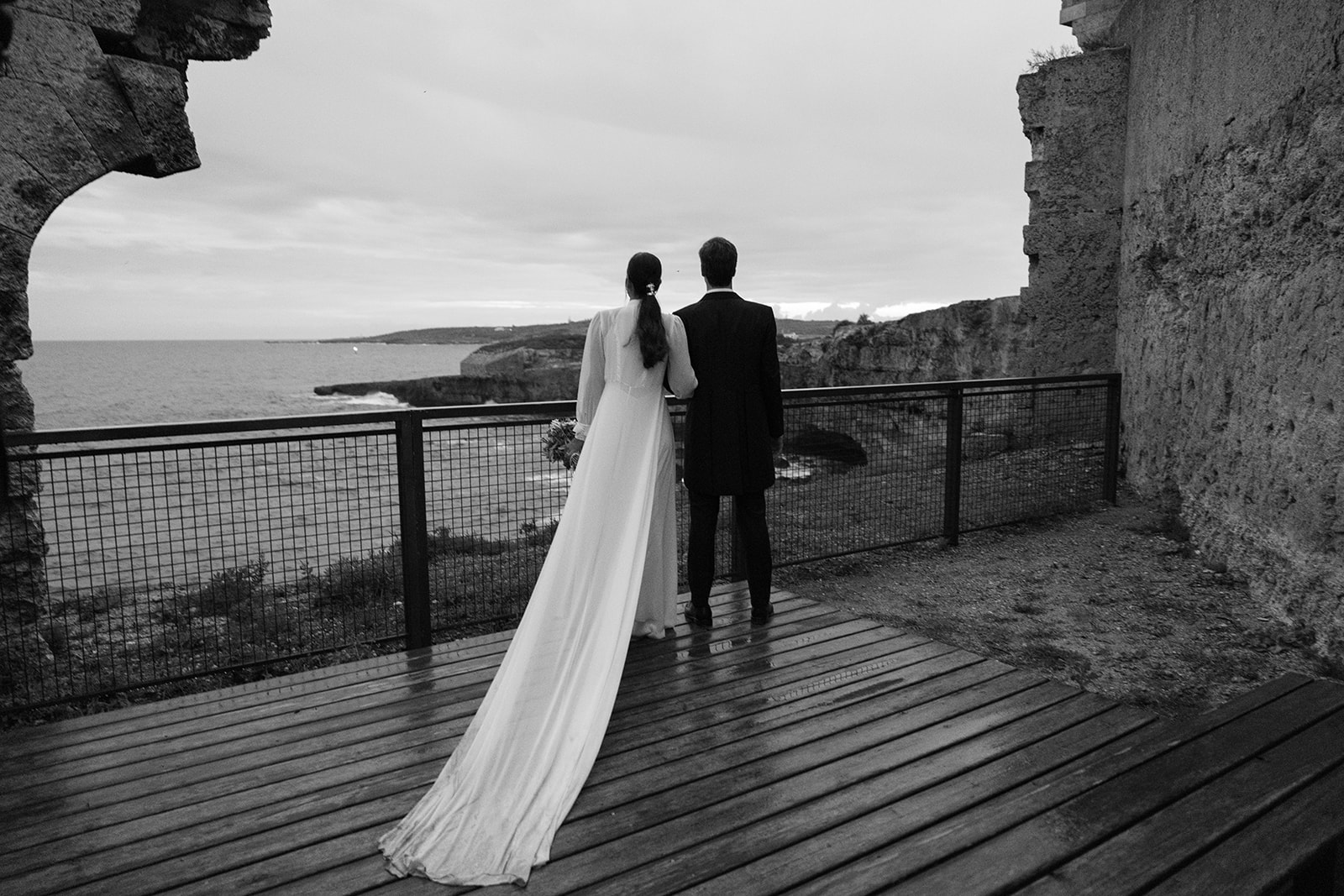 Bride and groom standing on the cliff at La Mola, Menorca, overlooking the sea — captured by Menorca wedding photographer Sus Pons.