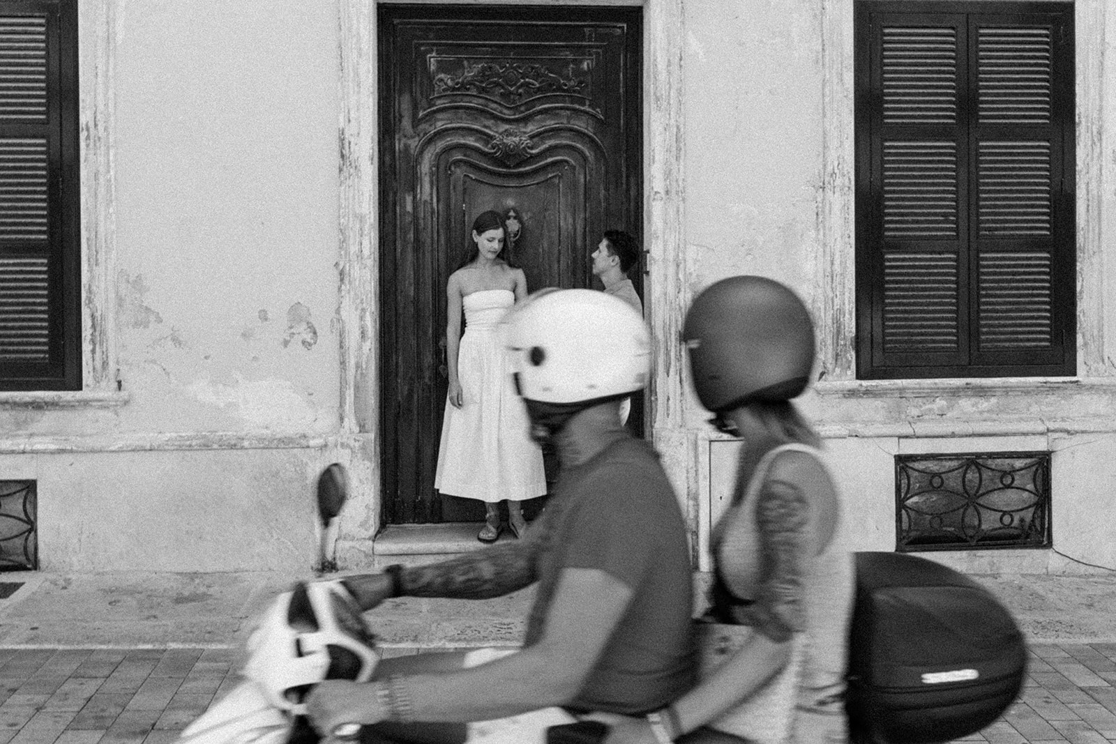 Bride in a minimalist white dress stands in front of an antique wooden door in Menorca while a couple on a scooter passes by — candid and cinematic street moment captured by Menorca wedding photographer Sus Pons.