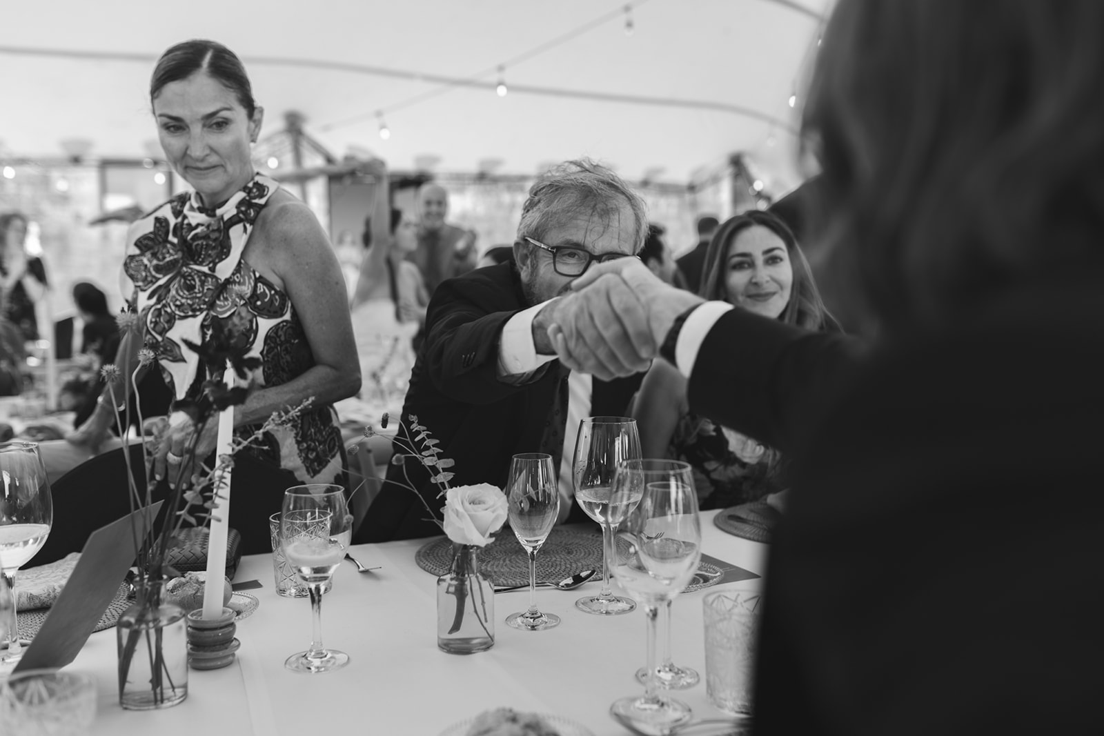 Candid wedding moment during dinner, guests shaking hands and smiling at the table, black and white photo by Sus Pons