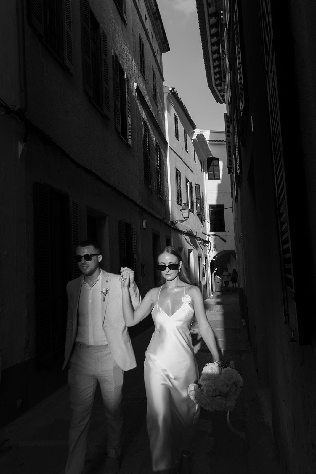 Bride and groom walking through the narrow sunlit streets of Ciutadella, Menorca, after their wedding ceremony.