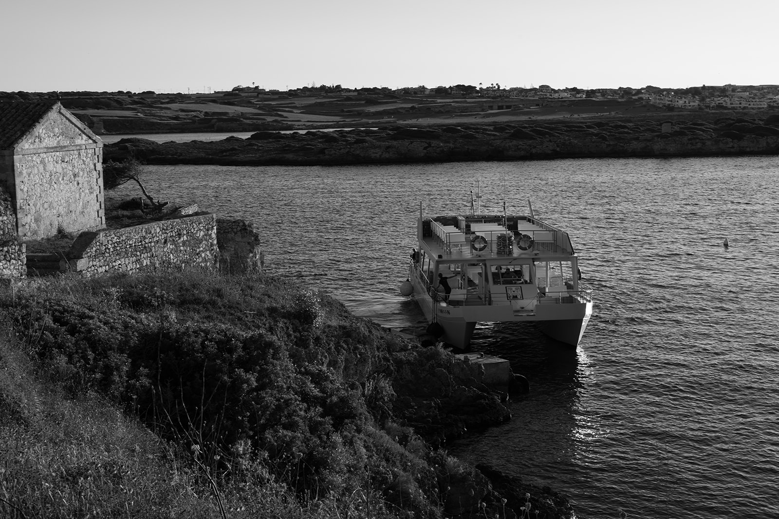 Black and white photo of a boat docked beside a stone building on the coast of Menorca, captured by Sus Pons